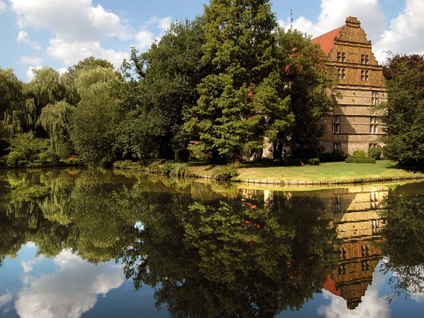 Wasserschloß Holtfeld Historisches Haus mit rotem Dach und Bäumen spiegelt sich idyllisch in einem ruhigen Teich wider.
