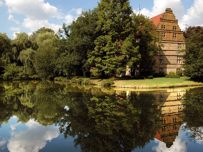 Wasserschloß Holtfeld Historisches Haus mit rotem Dach und Bäumen spiegelt sich idyllisch in einem ruhigen Teich wider.