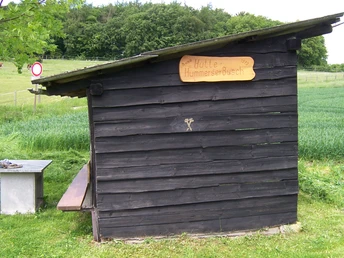 Holzhütte mit Flachdach auf Wiese vor Wald und Feldern; Tisch-Bank-Kombination daneben.