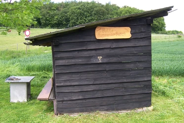 Schutzhütte Hummerser Busch Holzhütte mit Flachdach auf Wiese vor Wald und Feldern; Tisch-Bank-Kombination daneben.