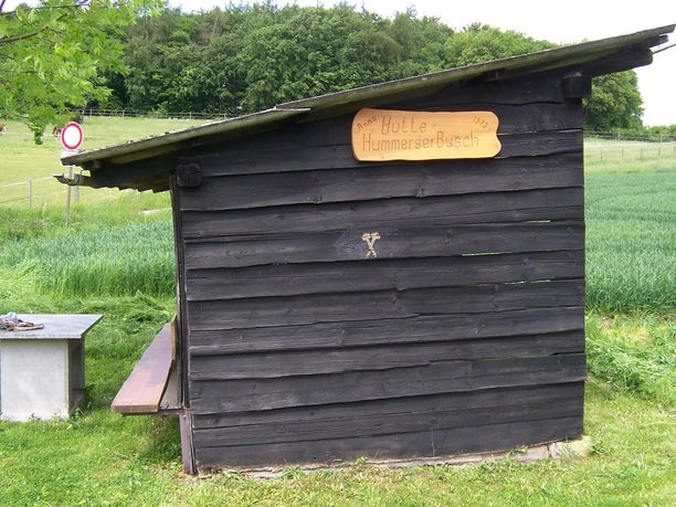 Schutzhütte Hummerser Busch Holzhütte mit Flachdach auf Wiese vor Wald und Feldern; Tisch-Bank-Kombination daneben.