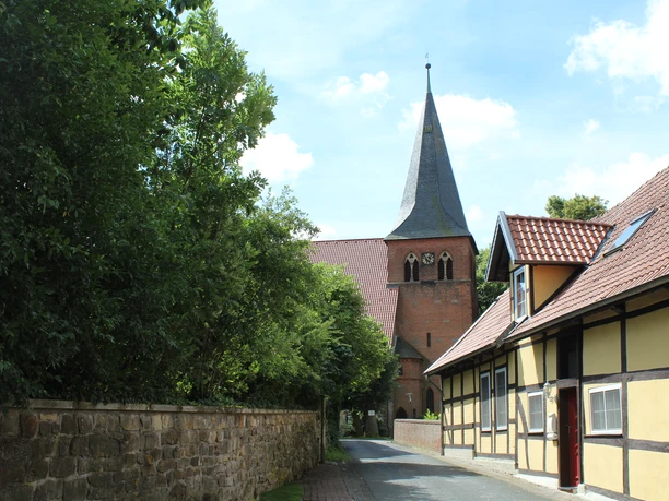Straße führt zu einer historischen Backsteinkirche mit spitzem Turm, flankiert von Fachwerkhäusern.