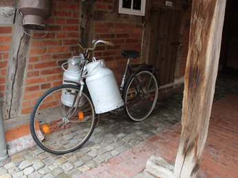 Ein vintage Fahrrad mit Milchkanne steht auf einem gepflasterten Hof an einer roten Backsteinwand.