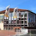 Kulturzentrum mit markanten Glasfassaden und Wimpel vorm Haupteingang, blauer Himmel im Hintergrund.