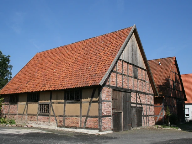 Fachwerkhaus mit rotem Ziegeldach und Holztür in Liebenau, umgeben von Bäumen und klarer Himmel.