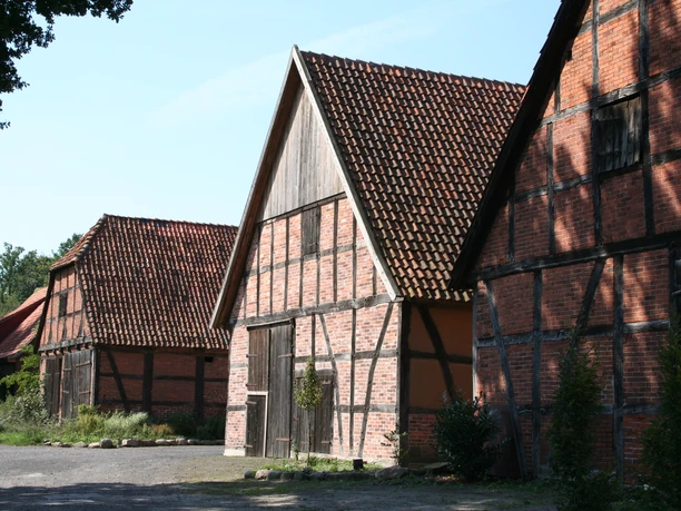 Drei historische Fachwerkscheunen mit roten Ziegeldächern stehen auf einer sonnigen, ruhigen Fläche.