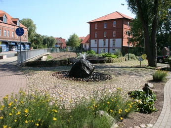 Natursteinbrunnen auf gepflastertem Platz vor historischer Wassermühle in Liebenau umgeben von Blumen.