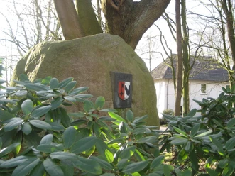Findling im Rathausgarten Großer Findling mit Wappen im grünen Rathausgarten, umgeben von Rhododendron und Bäumen.