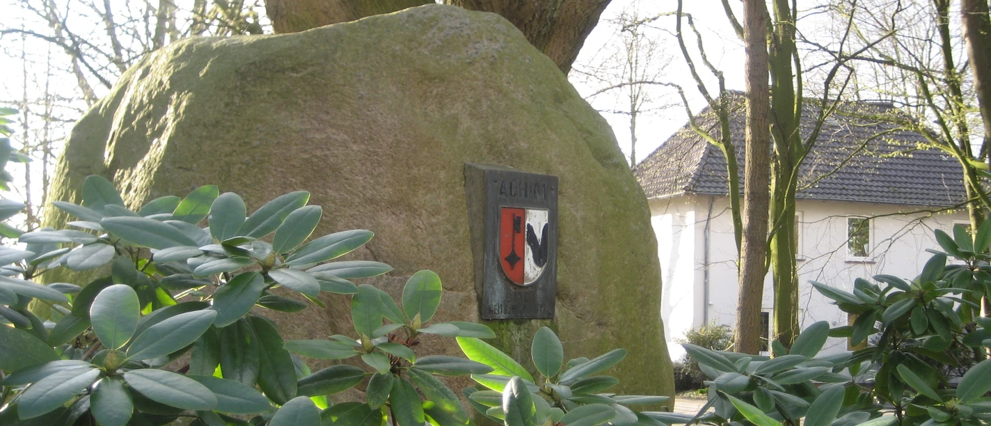Findling im Rathausgarten Großer Findling mit Wappen im grünen Rathausgarten, umgeben von Rhododendron und Bäumen.