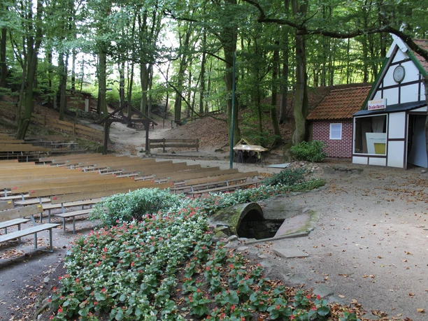 An open-air stage in a wooded setting with wooden spectator benches, flanked by a small building and flowering plants.