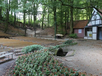 Freilichtbühne Marklohe Freiluftbühne in bewaldeter Umgebung mit Zuschauerbänken aus Holz, flankiert von einem kleinen Gebäude und blühenden Pflanzen.
