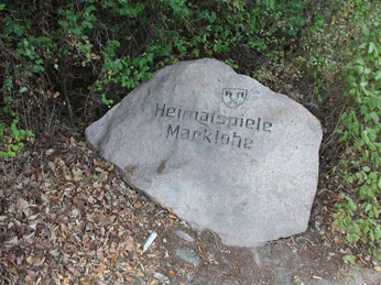 Freilichtbühne Marklohe Gedenkstein mit Inschrift "Heimspiele Marklohe" in naturbelassener Umgebung, umgeben von Blättern.