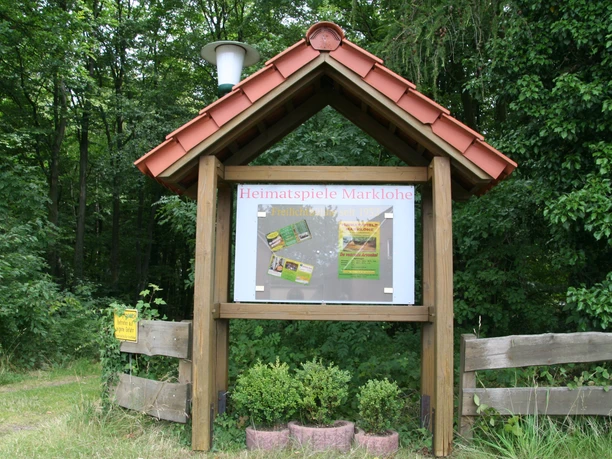 Freilichtbühne mit Infotafel und Werbeschildern, umgeben von üppigem Grün und hölzernem Zaun.
