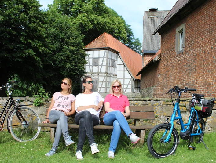 Drei Personen entspannen auf einer Bank vor dem historischen Kloster Schinna, umgeben von Fahrrädern.Three people relax on a bench in front of the historic Schinna Monastery, surrounded by bicycles.Tre personer slapper af på en bænk foran det historiske Schinna-kloster, omgivet af cykler.Drie mensen ontspannen op een bankje voor het historische klooster van Schinna, omringd door fietsen.