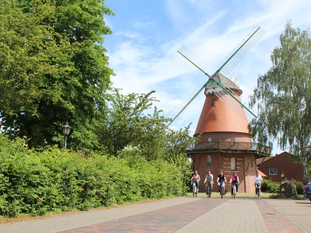A historic windmill in the countryside, frequented by cyclists, stands under a blue sky.