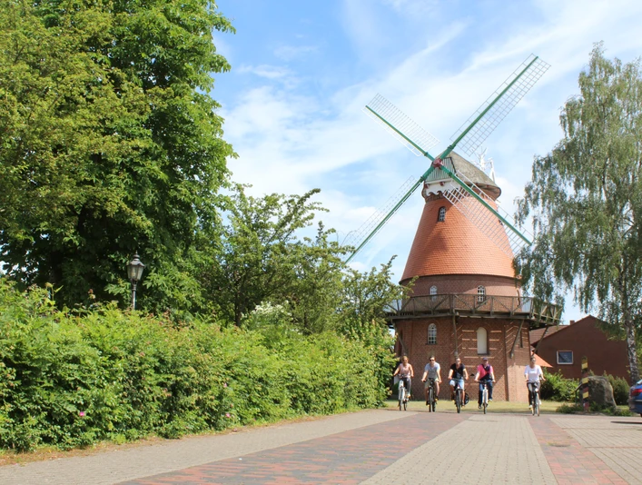 Eine historische Windmühle im Grünen, von Radfahrern besucht, steht unter blauem Himmel.