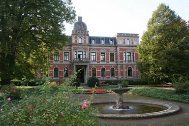 Historic castle with gardens and central fountain, surrounded by a well-kept park.