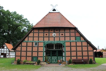 Brick half-timbered house with red tiled roof and green wooden ornamentation in front of meadow and trees.