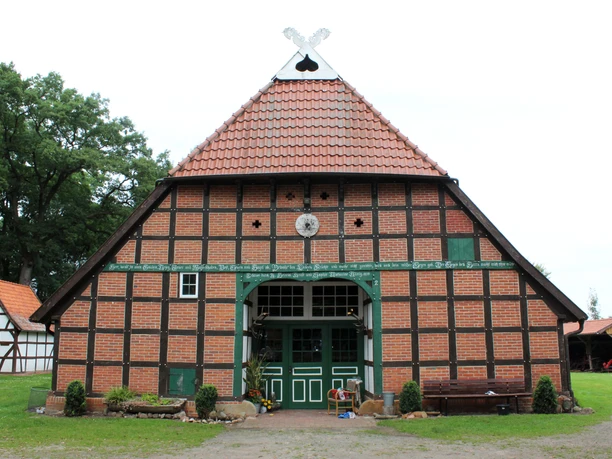 Brick half-timbered house with red tiled roof and green wooden ornamentation in front of meadow and trees.