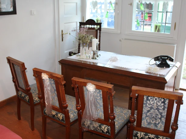 Drei verzierte Stühle stehen vor einem Holztisch in einem hellen Zimmer mit großen Fenstern.