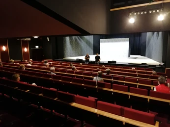 Leeres Theater mit rotem Gestühl und weiten Sitzreihen, auf der Bühne zwei Personen im Gespräch.