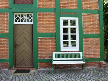 Historisches Backsteingebäude mit grünen Holzbalken und Bank daneben, umgeben von Natur.