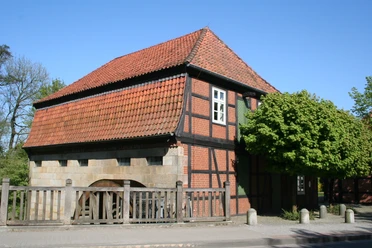 Fachwerkgebäude der Wassermühle Sudweyhe mit rotem Ziegeldach und grünem Laubbaum.