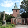 Backsteinkirche mit grünem Turmhelm, umgeben von Bäumen, daneben ein historischer Obelisk.