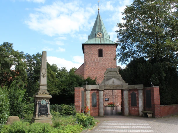 Backsteinkirche mit grünem Turmhelm, umgeben von Bäumen, daneben ein historischer Obelisk.