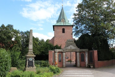 Marienkirche Leeste Backsteinkirche mit grünem Turmhelm, umgeben von Bäumen, daneben ein historischer Obelisk.