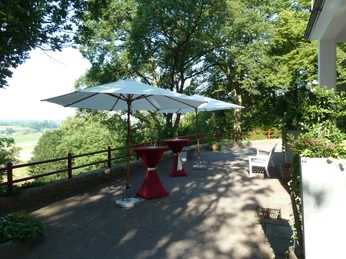 Terrasse mit Sonnenschirmen, Stehtischen in Rot und Blick auf grüne Landschaft unter Bäumen.