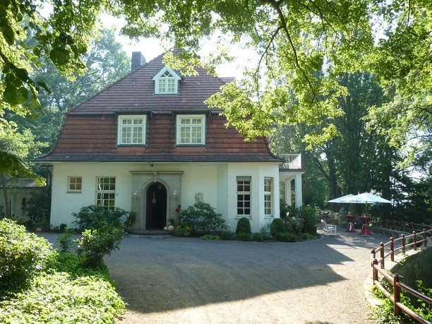 Haus Hünenburg Ein zweistöckiges Haus mit rotem Dach, umgeben von Bäumen, mit einem Schirmtisch im Garten.