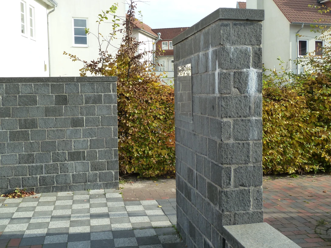 Gedenkstätte mit grauen Steinwänden und gepflastertem Boden, umgeben von Wohnhäusern und Büschen.