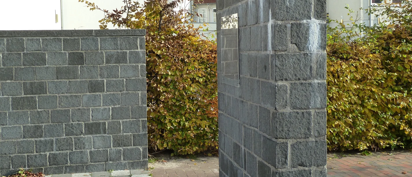 Jüdisches Mahnmal Gedenkstätte mit grauen Steinwänden und gepflastertem Boden, umgeben von Wohnhäusern und Büschen.