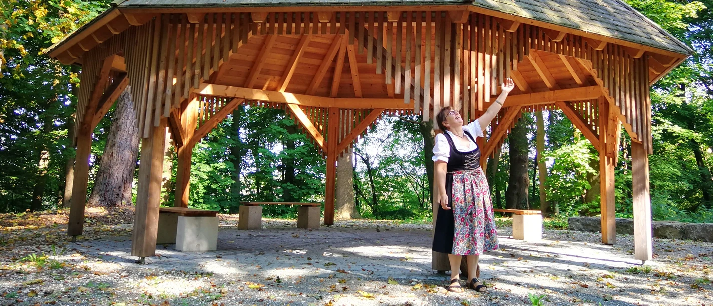 Gartenkunst. In Günzburg - Die Parkführung Holzpavillon im Wald mit Frau in Tracht, die das Dach bewundert, Sonnenlicht durch die Bäume.