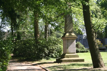 Steinernes Kriegerdenkmal von Bäumen umgeben, gedenkt an Konflikt von 1870/71; grüne Parkumgebung.