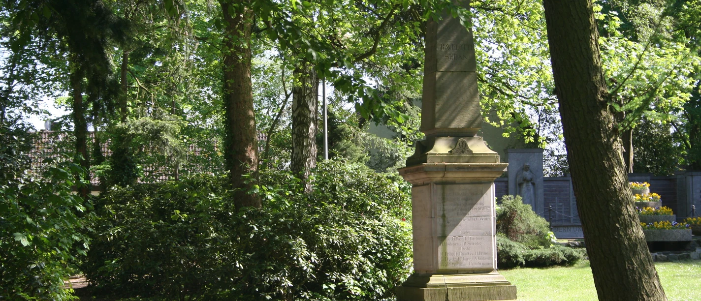 Steinernes Kriegerdenkmal von Bäumen umgeben, gedenkt an Konflikt von 1870/71; grüne Parkumgebung.