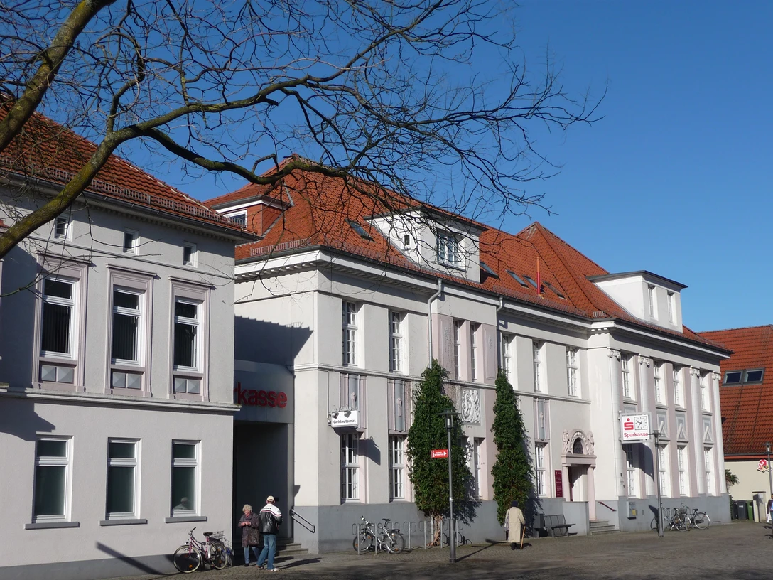 Historisches weißes Gebäude mit rotem Ziegeldach, Sitz der Sparkasse in Achim, Bäume im Vordergrund.