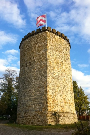 Ein runder mittelalterlicher Steinturm mit Zinnen und wehender rot-weißer Flagge vor blauem Himmel.