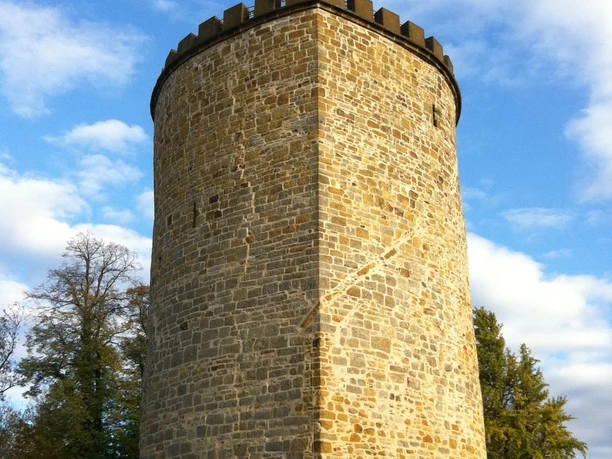 Ein runder mittelalterlicher Steinturm mit Zinnen und wehender rot-weißer Flagge vor blauem Himmel.