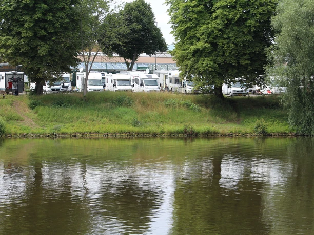 Wohnmobilstellplatz am Flussufer mit grünen Bäumen, grasbewachsenen Ufern und geparkten Fahrzeugen.