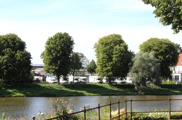Reisemobilstellplatz an Flussufer mit Wohnmobilen im Hintergrund und Bäumen im Vordergrund.