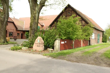 Unser kleiner Hof Wietzen Ein alter Bauernhof mit Backsteingebäuden, umgeben von grünen Bäumen und einem Weg aus Asphalt.