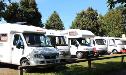 Wohnmobilstellplatz Reihen abgestellter Wohnmobile auf einem Stellplatz, umgeben von Bäumen bei klarem Wetter.