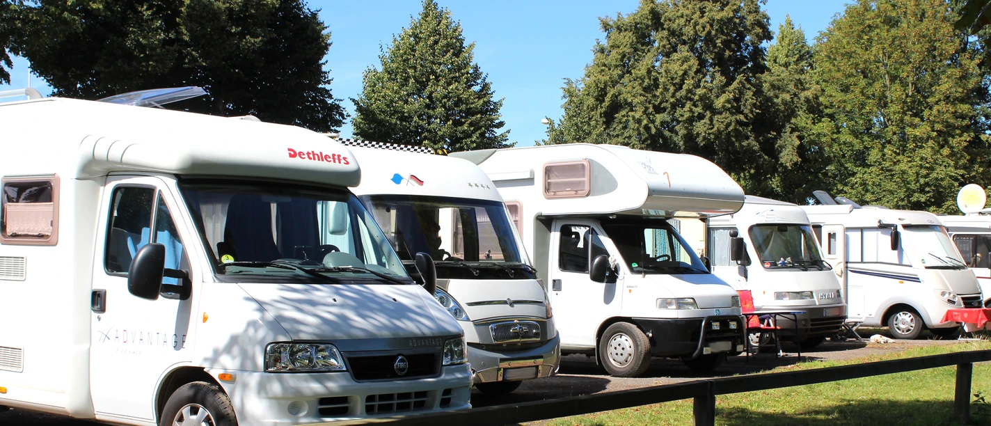 Wohnmobilstellplatz Reihen abgestellter Wohnmobile auf einem Stellplatz, umgeben von Bäumen bei klarem Wetter.