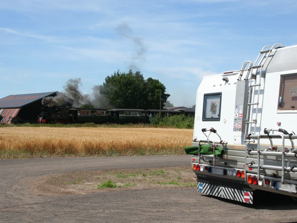 A motorhome next to a grain field, with a steaming locomotive and trees in the background near Bruchhausen-Vilsen.