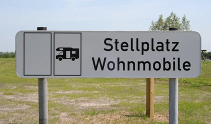 Wohnmobilstellplatz Schild für Wohnmobilstellplatz auf grünem Rasen vor blauem Himmel mit Bäumen im Hintergrund.