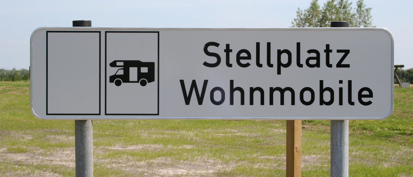 Wohnmobilstellplatz Schild für Wohnmobilstellplatz auf grünem Rasen vor blauem Himmel mit Bäumen im Hintergrund.