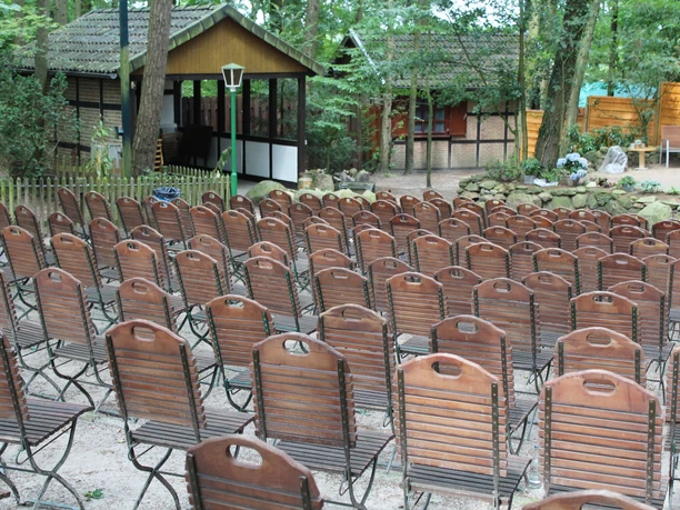 Freilichtbühne mit leeren Holzstühlen vor einer Kulisse aus Bäumen und rustikalen Gebäuden im Hintergrund.