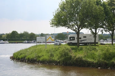 Wohnmobilstellplatz Marina Mehlbergen Wohnmobil auf begrüntem Platz an einer Marina, mit Bäumen und ruhigem Wasser im Hintergrund.